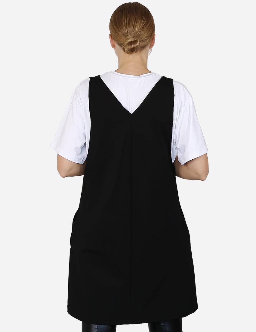 Back view of a woman wearing a stylish black pinafore dress over a white blouse, hands on hips.