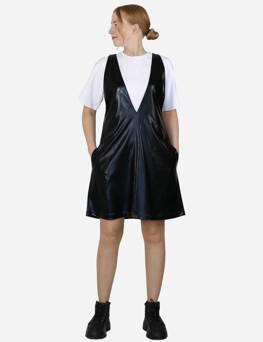 Back view of a woman standing with hands on hips wearing a black faux leather pinafore dress and white t-shirt.