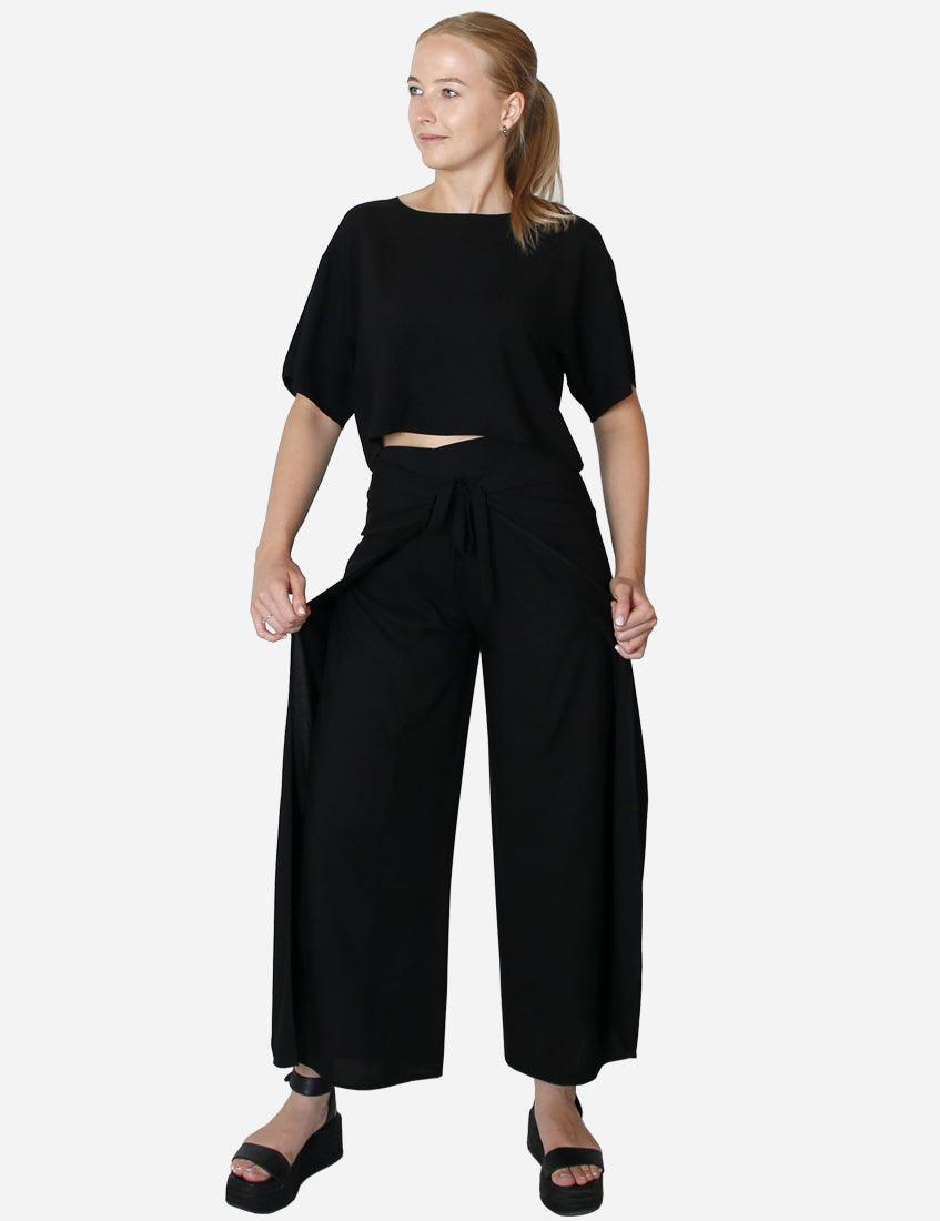 Woman in loose-fit black trousers and cropped top looking to the side with hands in pockets, showcasing a casual yet chic style.
