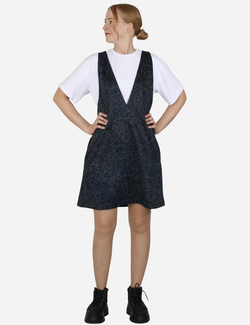 Woman in a blue patterned pinafore dress and white tee standing confidently with hands on hips.