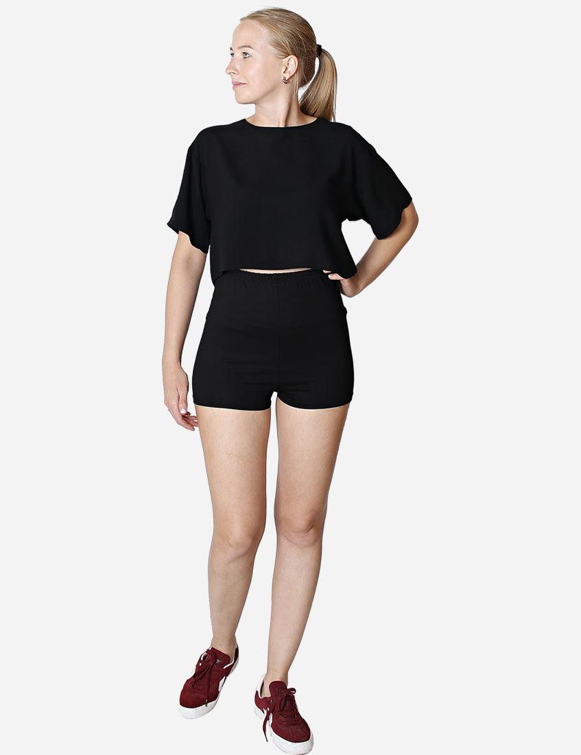 Elegant woman in high-waisted black shorts with a cropped top looking to the side, paired with maroon sneakers, isolated on a white background.