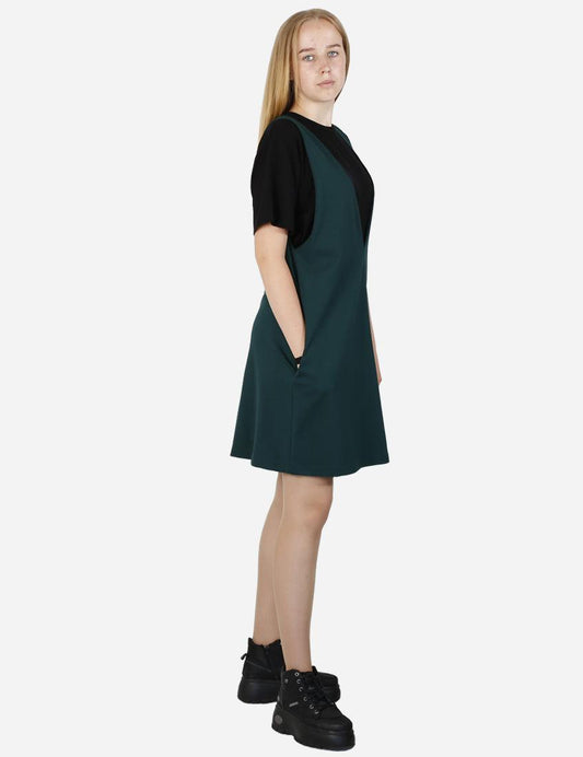 Side angle view of a woman wearing a pine green pinafore dress over a black t-shirt.