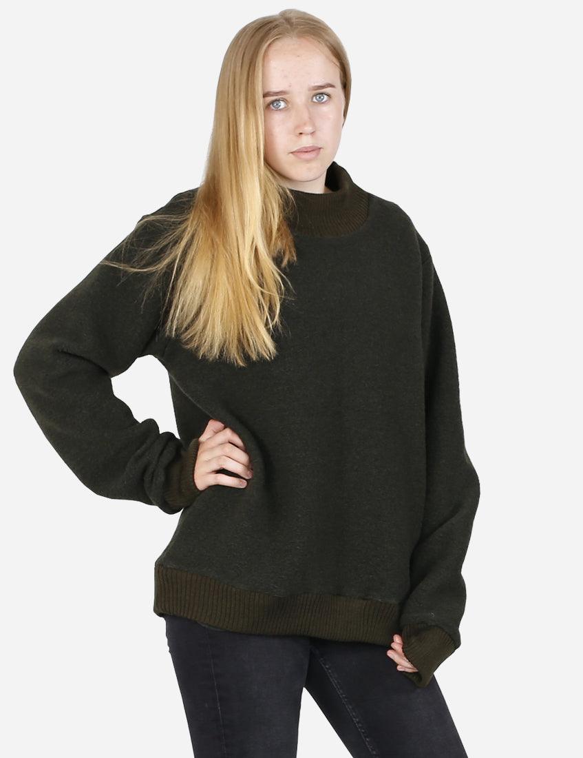 Young woman posing in a nature-inspired sage green woolen sweater with a ribbed turtleneck and cuffs, paired with slim-fit black jeans and lace-up boots.