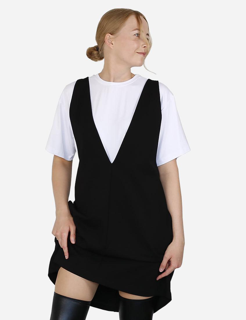 Side view of a woman in a black pinafore dress over a white blouse, casually standing.